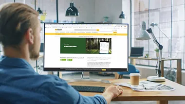 Over the Shoulder Shot: Man Uses Desktop Computer with Green Mock-up Screen while Sitting at His Desk. In the Background Stylish Modern Open Space Creative Business Office